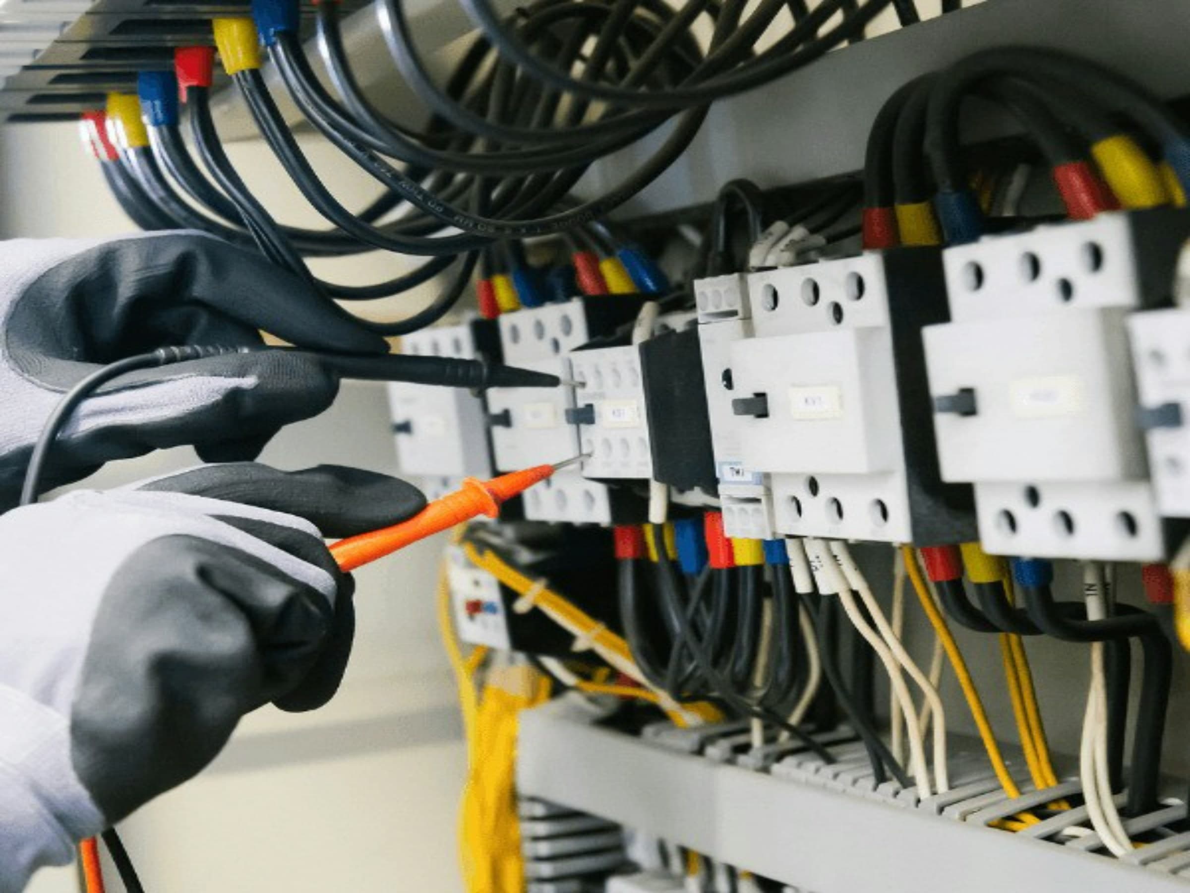 Gloved hand testing contactor terminals inside a control panel with a probe